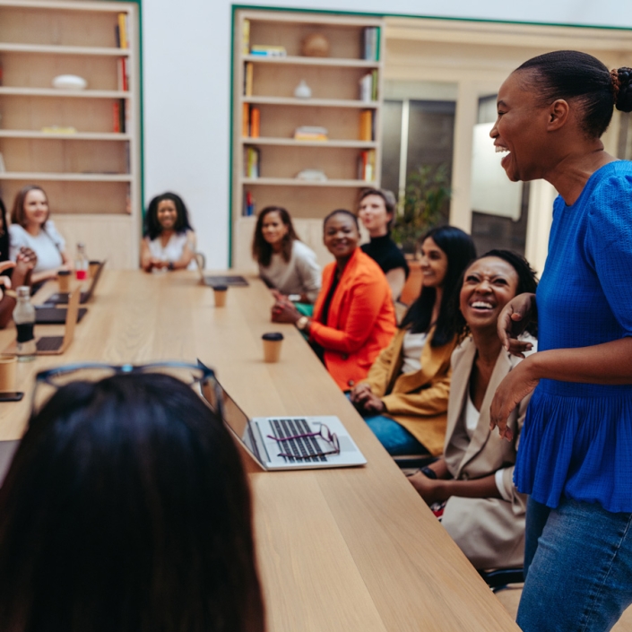 A large group of people sit around a conference table in a casual setting. A 30yo black woman is standing, in the foreground, speaking and laughing. The attendees are engaged and listening, smiling