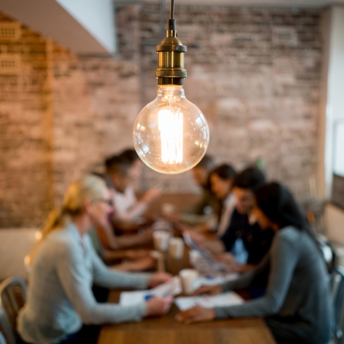 soft focus image of a diverse group of people sitting at a table, talking. Papers, laptops, and cups of coffee are in front of them. A modern light fixture featuring an Edison-style bulb is centered and in focus, with the working group behind/below.